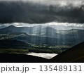 Sunlight breaks through storm clouds over the rolling hills and a lake in rural County Kerry, Ireland. The sunbeams illuminate the landscape during the daytime. 135489131