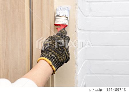 A woman's gloved hand paints a wall with white paint using a brush 135489748
