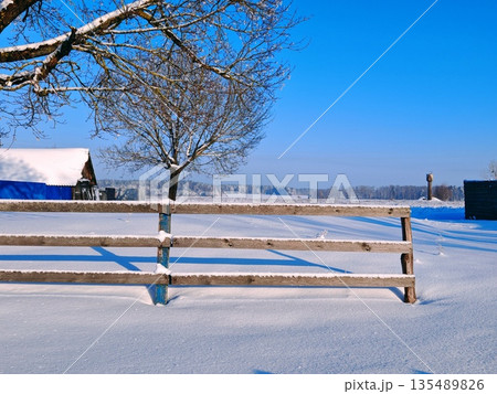 Snow Covered Wooden Fence Rural Winter. Rustic wooden fence covered in fresh snow with bare tree and distant snowy fields under bright blue sky. Cozy winter countryside scene and rural charm. 135489826