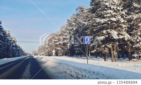 Snowy Forest Road with Pine Trees. Winter road through snow-covered pine forest under clear blue sky, road sign visible. Scenic winter drive, nature and seasonal travel concept. 135489894