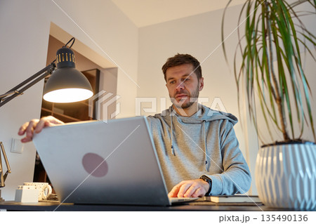 Man standing at home office desk using laptop under desk lamp in apartment interior 135490136