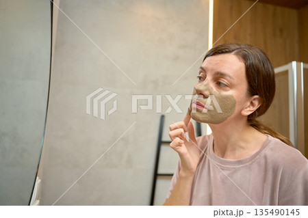Woman standing in bathroom and applying clay face mask while looking at mirror 135490145