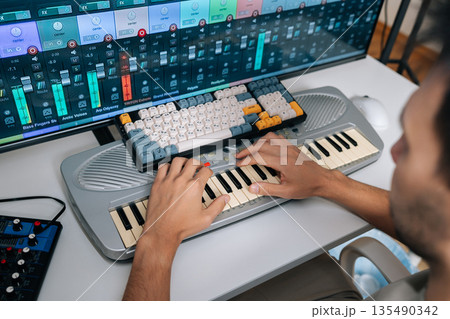View from shoulder of audio engineer creating new music in professional studio setup, inputting melodies on keyboard synthesizer while adjusting sound levels and effects on software interface closeup. 135490342