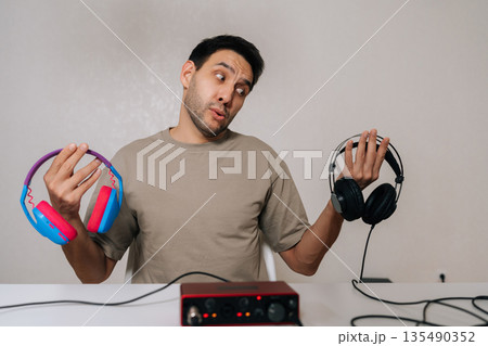 Portrait of confused excited man weighing colorful expressive headphones against black professional studio pair tied to audio interface, pondering audio quality, style, creative vs technical choice. 135490352