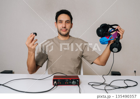 Portrait of young pensive man comparing studio headphones and in-ear earbuds sitting at white desk with audio interface, evaluating sound quality and gear choices for home studio production. 135490357