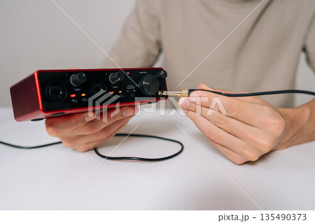 Person hands connecting audio jack cable into red usb audio interface, preparing for home music production, recording, and sound engineering in professional studio setup. 135490373