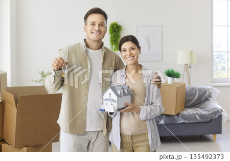 Happy couple holding wooden house model and new keys on moving day at home Happy couple holding wooden house model and new keys on moving day at home 135492373