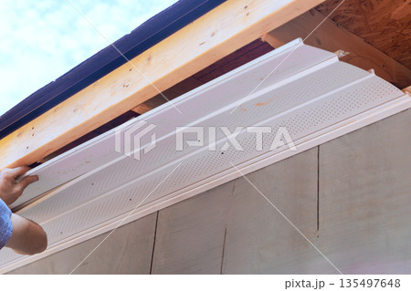 Worker installs white soffit panels on building while standing on ladder 135497648