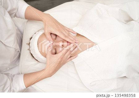 Young caucasian female receiving relaxing facial massage at spa in white robe Young caucasian female receiving relaxing facial massage at spa in white robe 135498358
