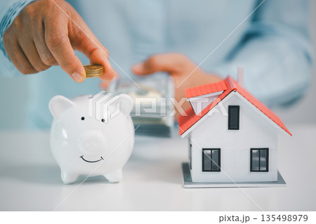 Hand holds coins over a white piggy bank beside a house model while cash is counted in the background, symbolizing mortgage down payment savings, real estate budgeting and smart finance. Hand holds coins over a white piggy bank beside a house model while cash is counted in the background, symbolizing mortgage down payment savings, real estate budgeting and smart finance. 135498979