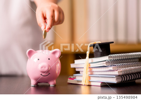 Hand puts a coin into pink piggy bank beside graduation cap and stacked notebooks on desk, symbolizing education savings, tuition budgeting, scholarship plans and smart student finance. Hand puts a coin into pink piggy bank beside graduation cap and stacked notebooks on desk, symbolizing education savings, tuition budgeting, scholarship plans and smart student finance. 135498984