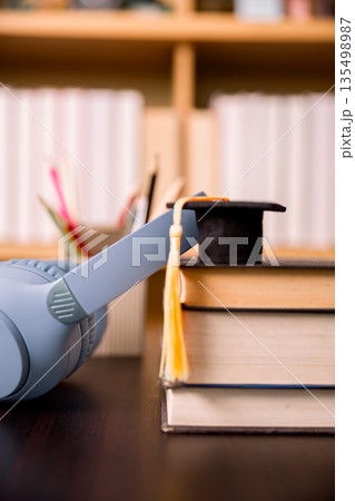 Gray headphones beside stacked textbooks with graduation cap and tassel on desk, blurred library background. Concept for online learning, audiobooks, study focus, education success. 135498987