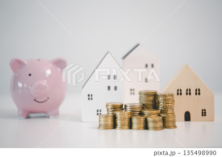 Wooden house model, coin stacks and pink piggy bank on white background, representing home savings, mortgage down payment, real estate budget planning and financial security goals. 135498990