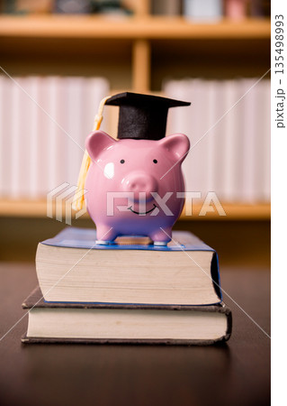 Pink piggy bank with graduation cap on stacked textbooks in front of blurred bookshelf, symbolizing education savings, tuition funding, budgeting, scholarships and smart financial planning. 135498993