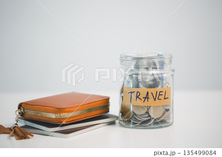 Glass jar labeled TRAVEL filled with coins and cash beside a leather wallet and notebooks on white background, symbolizing vacation savings, budget planning, travel fund goals and expenses. Glass jar labeled TRAVEL filled with coins and cash beside a leather wallet and notebooks on white background, symbolizing vacation savings, budget planning, travel fund goals and expenses. 135499084