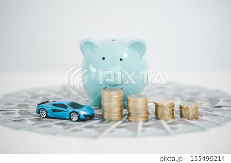 Blue piggy bank behind stacks of coins and a toy car on scattered dollar bills, representing car savings, down payment, auto loan budgeting, insurance costs and smart finance planning. Blue piggy bank behind stacks of coins and a toy car on scattered dollar bills, representing car savings, down payment, auto loan budgeting, insurance costs and smart finance planning. 135499214