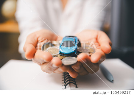 Hands hold a toy car on a pile of coins above a notebook and pen, symbolizing car savings, down payment, auto loan budgeting, insurance costs and smart finance planning for buyers and future goals. 135499217