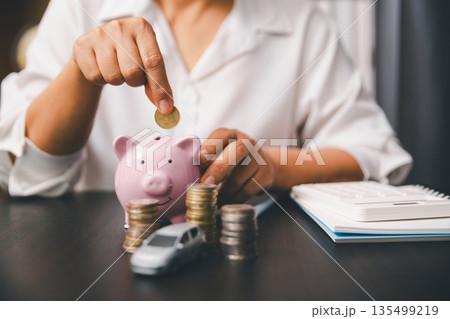 Hand drops a coin into a pink piggy bank beside coin stacks, a toy car and calculator on desk, showing car savings, down payment, auto loan budgeting, insurance costs and finance planning. Hand drops a coin into a pink piggy bank beside coin stacks, a toy car and calculator on desk, showing car savings, down payment, auto loan budgeting, insurance costs and finance planning. 135499219