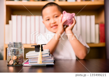 Child holding a piggy bank behind desk with coin jar, cash and graduation cap on notebooks, representing education savings, tuition planning, family budgeting and future financial goals. 135499221