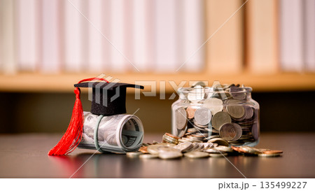 Graduation cap on a rolled banknote beside a glass jar of coins on a desk, symbolizing tuition savings, student budget planning, scholarships and financial goals for education funding and success. Graduation cap on a rolled banknote beside a glass jar of coins on a desk, symbolizing tuition savings, student budget planning, scholarships and financial goals for education funding and success. 135499227