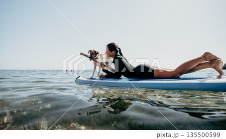 Woman Dog Paddleboarding Ocean: A woman and her dog paddleboard in the ocean on a sunny day. Woman Dog Paddleboarding Ocean: A woman and her dog paddleboard in the ocean on a sunny day. 135500599