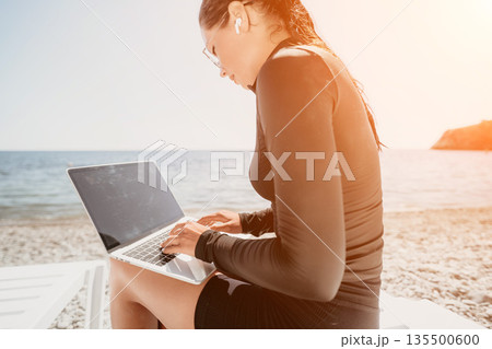 Woman Beach Laptop Work: Woman working on her laptop while sitting on the beach on a sunny day. 135500600