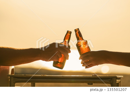 Two cold, steamed beer bottles in their hands clink glasses in close-up on the beach, against the background of the setting sun at sunset, sand and sea. Vacation and travel thirst quenching. High 135501207