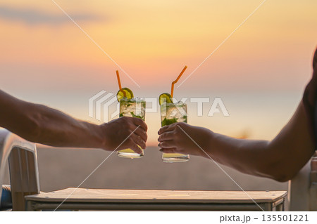 Lovers hands hold mojito cocktail glasses against the background of a sea sunset. They celebrate Valentine Day on a beach vacation or honeymoon and say Cheers High quality photo 135501221