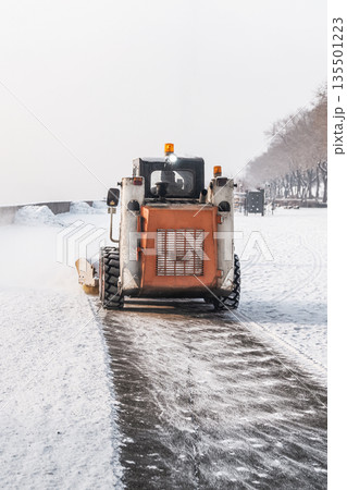 A mini loader that removes snow on a snowy city embankment or pedestrian sidewalk after winter snowfall. Cleaning urban infrastructure from snow and debris in winter by municipal services of the city A mini loader that removes snow on a snowy city embankment or pedestrian sidewalk after winter snowfall. Cleaning urban infrastructure from snow and debris in winter by municipal services of the city 135501223