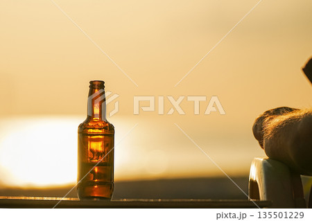 A bottle of beer on the table during sunset at sea is illuminated by the orange sun, the concept of a holiday at the resort. Relaxing and enjoying a drink in the evening and observing nature. High A bottle of beer on the table during sunset at sea is illuminated by the orange sun, the concept of a holiday at the resort. Relaxing and enjoying a drink in the evening and observing nature. High 135501229
