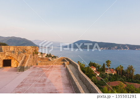 A historic stone terrace overlooks a tranquil bay with distant hills and a coastal town 135501291