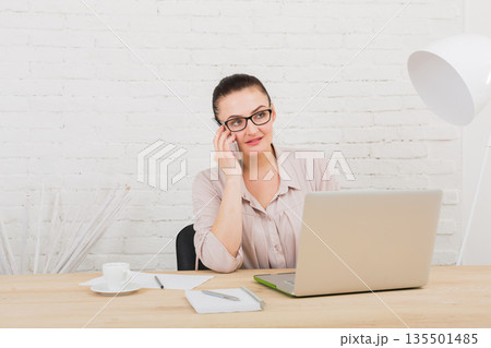 Middle-aged business woman in office with smartphone. Female using laptop computer and talking on mobile phone at workplace, copy space. Communication concept Middle-aged business woman in office with smartphone. Female using laptop computer and talking on mobile phone at workplace, copy space. Communication concept 135501485