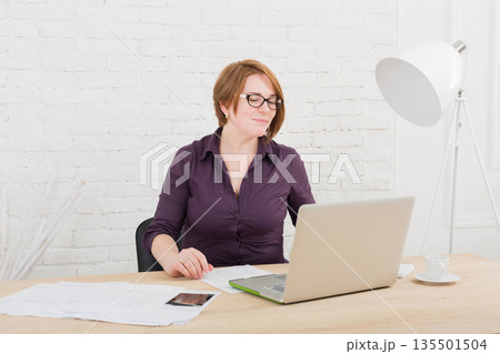 Middle-aged Woman in the office. Female architect using laptop computer and smartphone. Office worker. Redhead caucasian woman in protective glasses with computer. High-key, soft tone. Middle-aged Woman in the office. Female architect using laptop computer and smartphone. Office worker. Redhead caucasian woman in protective glasses with computer. High-key, soft tone. 135501504