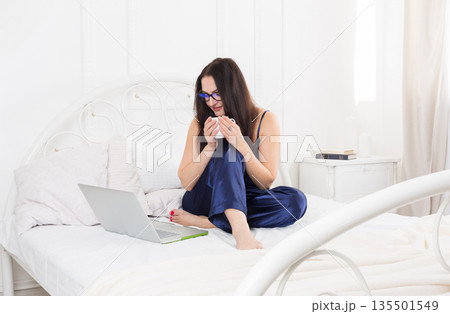 Morning coffee and news. Middle aged woman with laptop in bed. Beautiful woman in blue nightwear surfing internet at her computer. High key. 135501549