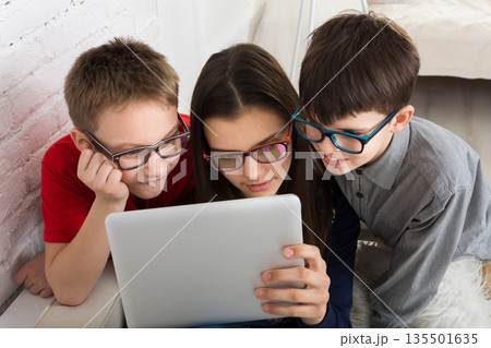 Children computer games, social networks and media addiction concept. Group of kids in eye glasses look into tablet. Girl and boys with tablet. Children computer games, social networks and media addiction concept. Group of kids in eye glasses look into tablet. Girl and boys with tablet. 135501635