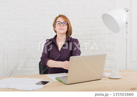 Middle-aged woman in office. Female architect using laptop and smartphone. Redhead caucasian woman in protective glasses with computer. Middle-aged woman in office. Female architect using laptop and smartphone. Redhead caucasian woman in protective glasses with computer. 135501660