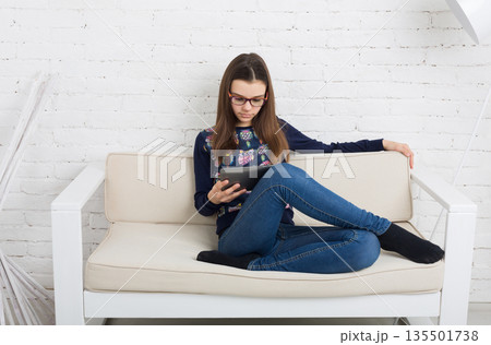 Teenage girl in eye glasses look into tablet, sitting on sofa. Children computer games, social networks and media addiction concept. Communication technologies, copy space 135501738