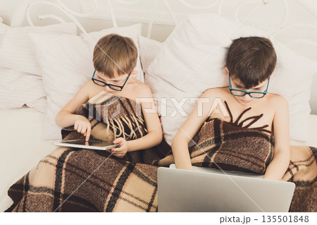 Boys in parents' bed at morning with laptop and tablet. Brothers play computer games. Siblings and gadgets. Children in glasses. 135501848