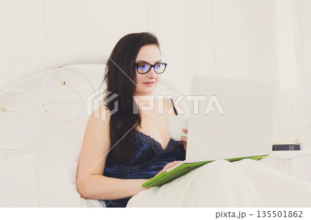 Sunday morning in bed. Middle aged woman in blue nightwear surfing internet at her laptop computer, copy space 135501862