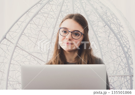 Beautiful smiling woman in eyeglasses in fashionable armchair with laptop. Girl with pc in modern interior. Stylish portrait of freelancer, work at home concept Beautiful smiling woman in eyeglasses in fashionable armchair with laptop. Girl with pc in modern interior. Stylish portrait of freelancer, work at home concept 135501866
