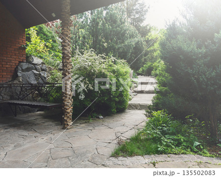 Beautiful landscape design, garden path with staircase in stone tiles, evergreen bushes and shrubs in sunlight. Bench in beautiful park. Summer yard design. 135502083