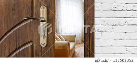 Half opened door handle closeup, entrance to a living room. View of sofa. Welcome, privacy concept. Door lock with keys, white brick wall, modern interior design. 135502352