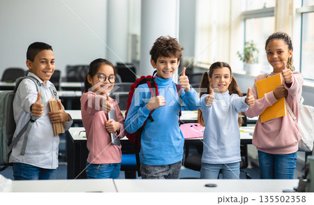 Education Concept. Diverse group of happy smiling international classmates standing in line with notebooks and backpacks in classroom and showing thumbs up sign gesture, enjoying studying at school Education Concept. Diverse group of happy smiling international classmates standing in line with notebooks and backpacks in classroom and showing thumbs up sign gesture, enjoying studying at school 135502358