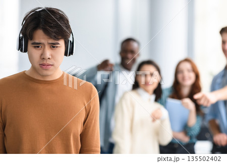 Group Conflict. Sad asian guy in headphones standing away alone, suffering humiliation and public disgrace, crowd of bad people bullying pointing fingers at victim, laughing. Racial Discrimination Group Conflict. Sad asian guy in headphones standing away alone, suffering humiliation and public disgrace, crowd of bad people bullying pointing fingers at victim, laughing. Racial Discrimination 135502402
