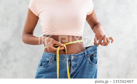 Closeup of black woman in old big jeans measuring her waist with tape, showing results of slimming diet or liposuction, grey background. Young lady promoting healthy nutrition for weight loss Closeup of black woman in old big jeans measuring her waist with tape, showing results of slimming diet or liposuction, grey background. Young lady promoting healthy nutrition for weight loss 135502407