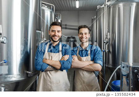Happy employees at favorite job, small business and male work. Young cheerful men workers in aprons with crossed arms look at camera in interior of factory with large boilers, free space Happy employees at favorite job, small business and male work. Young cheerful men workers in aprons with crossed arms look at camera in interior of factory with large boilers, free space 135502434