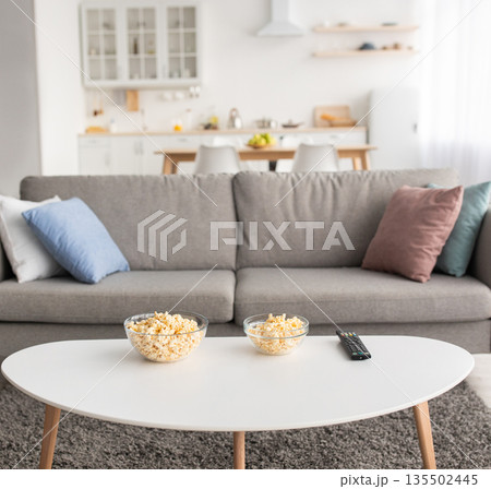 Movie night or online sports match at home during covid lockdown. Table with popcorn and remote control, sofa with pillows in living room, on kitchen interior background, nobody, flat lay, copy space Movie night or online sports match at home during covid lockdown. Table with popcorn and remote control, sofa with pillows in living room, on kitchen interior background, nobody, flat lay, copy space 135502445