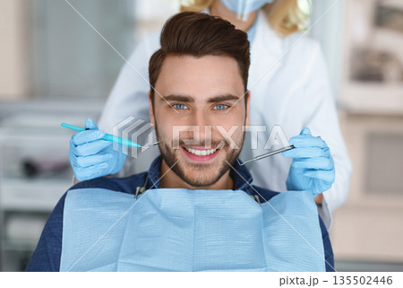 Smiling young bearded man sitting in dental chair, visiting modern dental clinic, having regular checkup, unrecognizable woman dentist holding dental tools next to male patient mouth 135502446