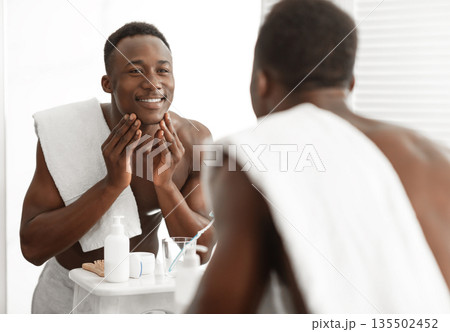 Happy Black Man Touching Smooth Face Skin After Shaving Standing Near Mirror In Bathroom At Home. Male Facial Skincare, Morning Beauty Routine And Self-Care Cosmetics Concept 135502452