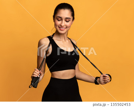 Cardio Workout. Portrait Of Confident Smiling Young Sportswoman In Black Bra Holding Skipping Rope Around Neck, Posing At Studio And Looking Down, Isolated On Orange Background, Banner Cardio Workout. Portrait Of Confident Smiling Young Sportswoman In Black Bra Holding Skipping Rope Around Neck, Posing At Studio And Looking Down, Isolated On Orange Background, Banner 135502453
